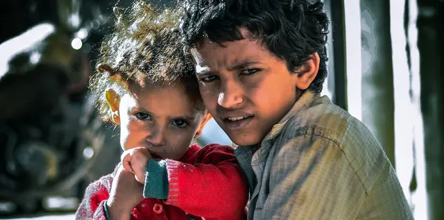 An older boy holds a younger child dressed in red close to him; both look towards the camera.