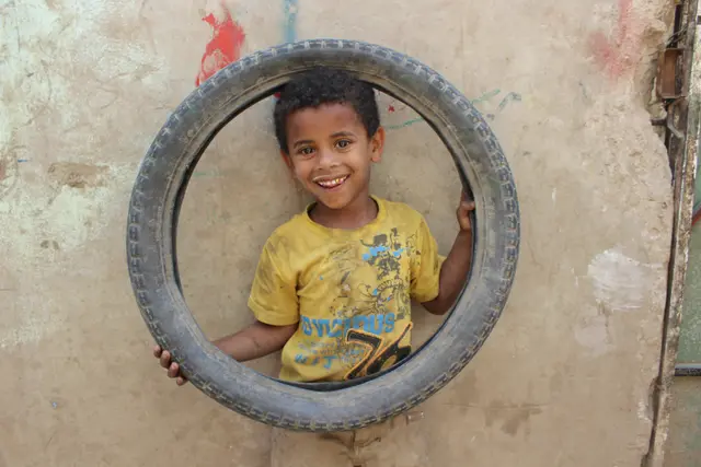 A smiling boy in a yellow T-shirt looks out through the centre of a tyre held against a wall.