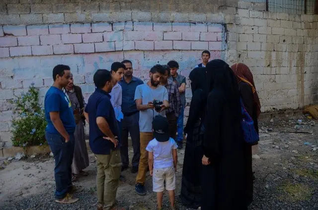 A group of people, some holding cameras, gathers in front of a whitewashed brick wall.