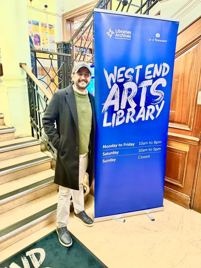 A man in a cap and long coat stands on a staircase beside a blue West End Arts Library banner.