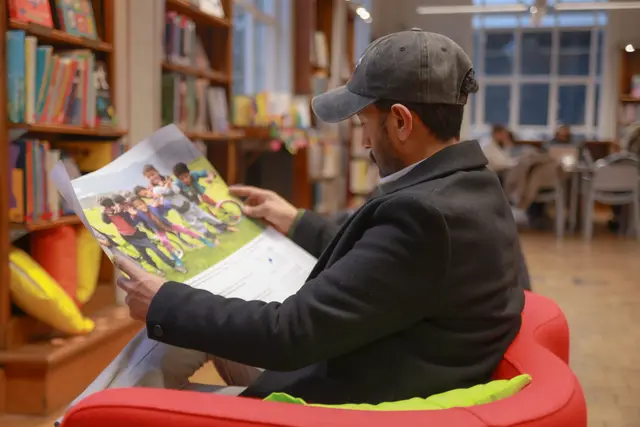 A man in a cap sits in a red library chair, reading an exhibition leaflet.