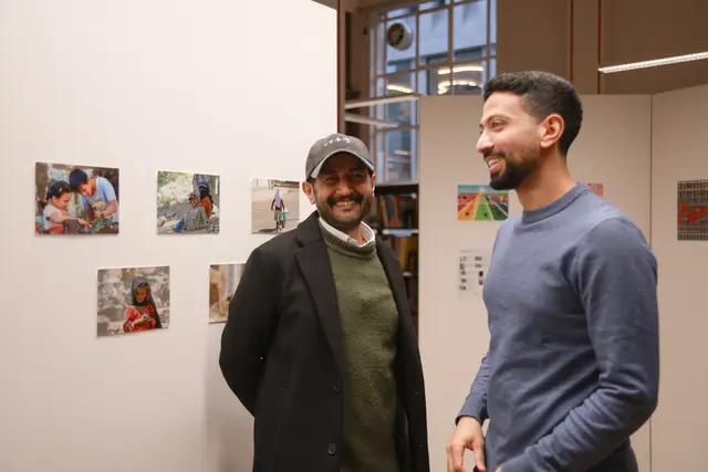 Two men smile in conversation in front of a wall of photographs.
