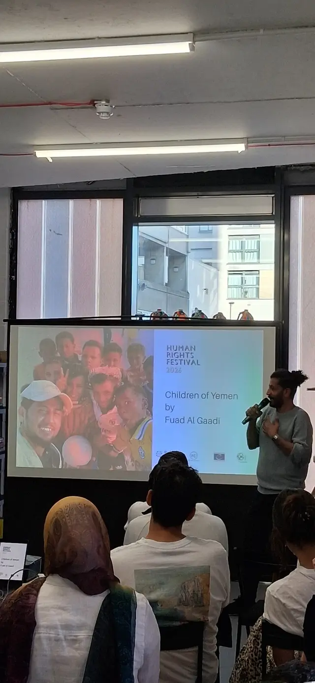 متحدث يحمل ميكروفونًا يقف بجانب شاشة كُتب عليها «Children of Yemen by Fuad Al Gaadi» ضمن مهرجان حقوق الإنسان 2026، أمام جمهور جالس.
