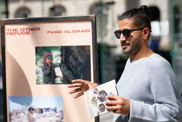 رجل بنظارة شمسية يحمل صورًا بجانب لوحة عنوانها «The Other Refuge — Fuad Al-Gaadi».