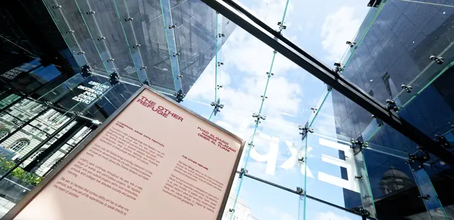 An exhibition text panel for The Other Refuge stands in a glass atrium.