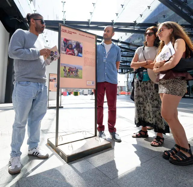 A man talks with visitors beside a freestanding exhibition panel in the atrium.