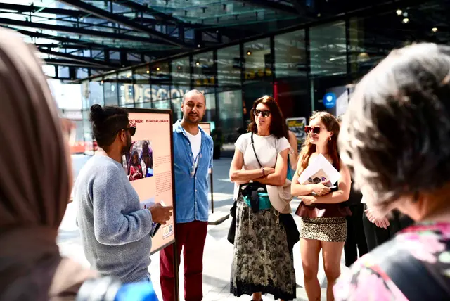 A man speaks to listeners beside an exhibition panel in the atrium.