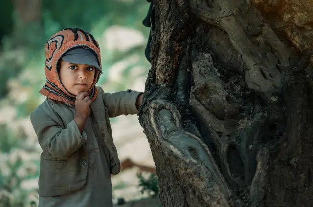 A young child in a knitted hat and coat stands beside a gnarled tree trunk, one hand raised to the chin.